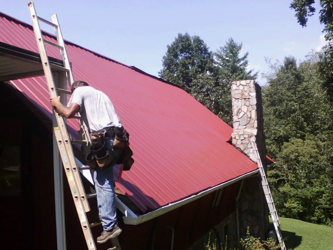 Roofer on a ladder beside a red metal roof and stone chimney — Soffit & Fascia Repair by Roofing Brothers in Wayne